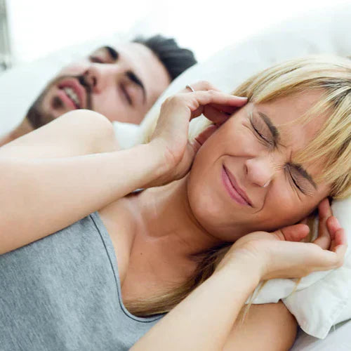Woman closes her ears with her fingers as her male partner snores behind her. Mouth Tape Reduces Snoring